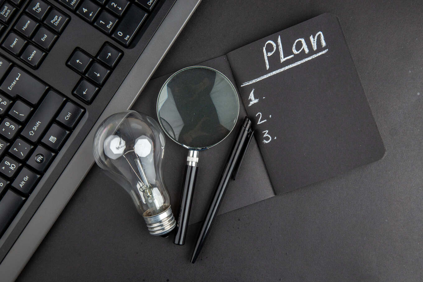 top-view-plan-written-black-notepad-lupa-keyboard-light-bulb-pen-black-table